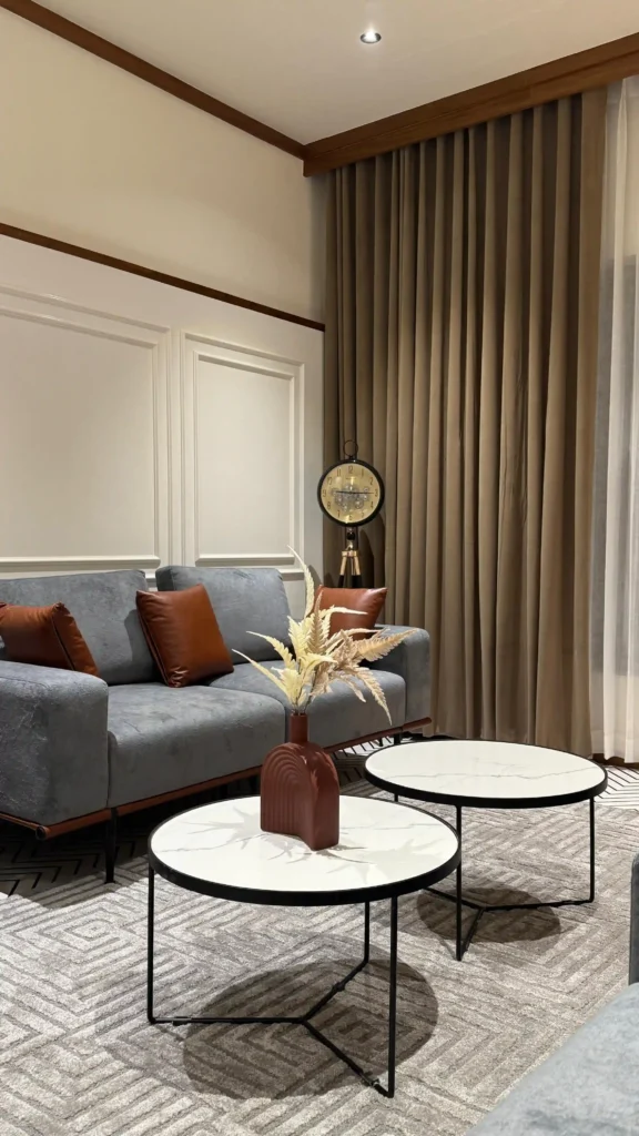 Modern living room with a gray sofa, brown accent pillows, round white coffee tables, a floor clock, and beige curtains